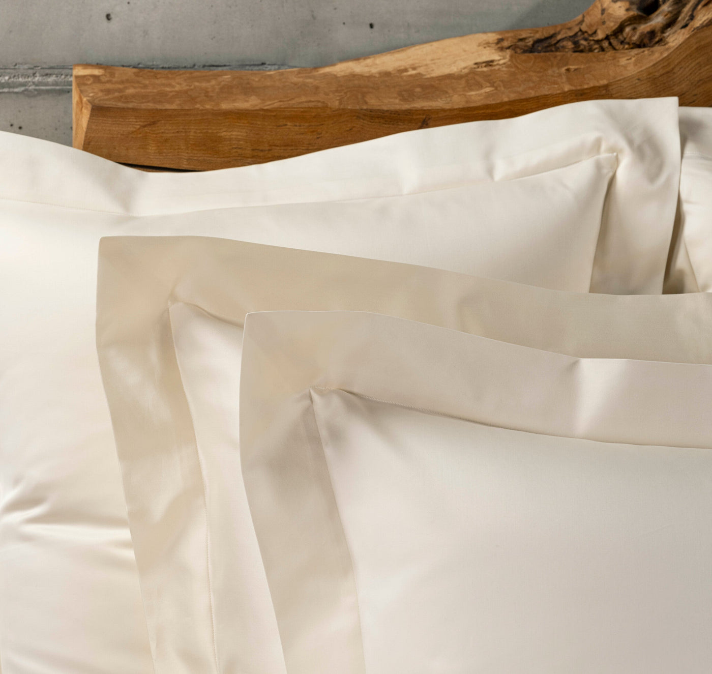Close-up of layered white pillowcases with a slight sheen and embroidered details, set against a rustic wooden headboard. featuring Capela Pillowcase by Celso de Lemos available at Linen Society.