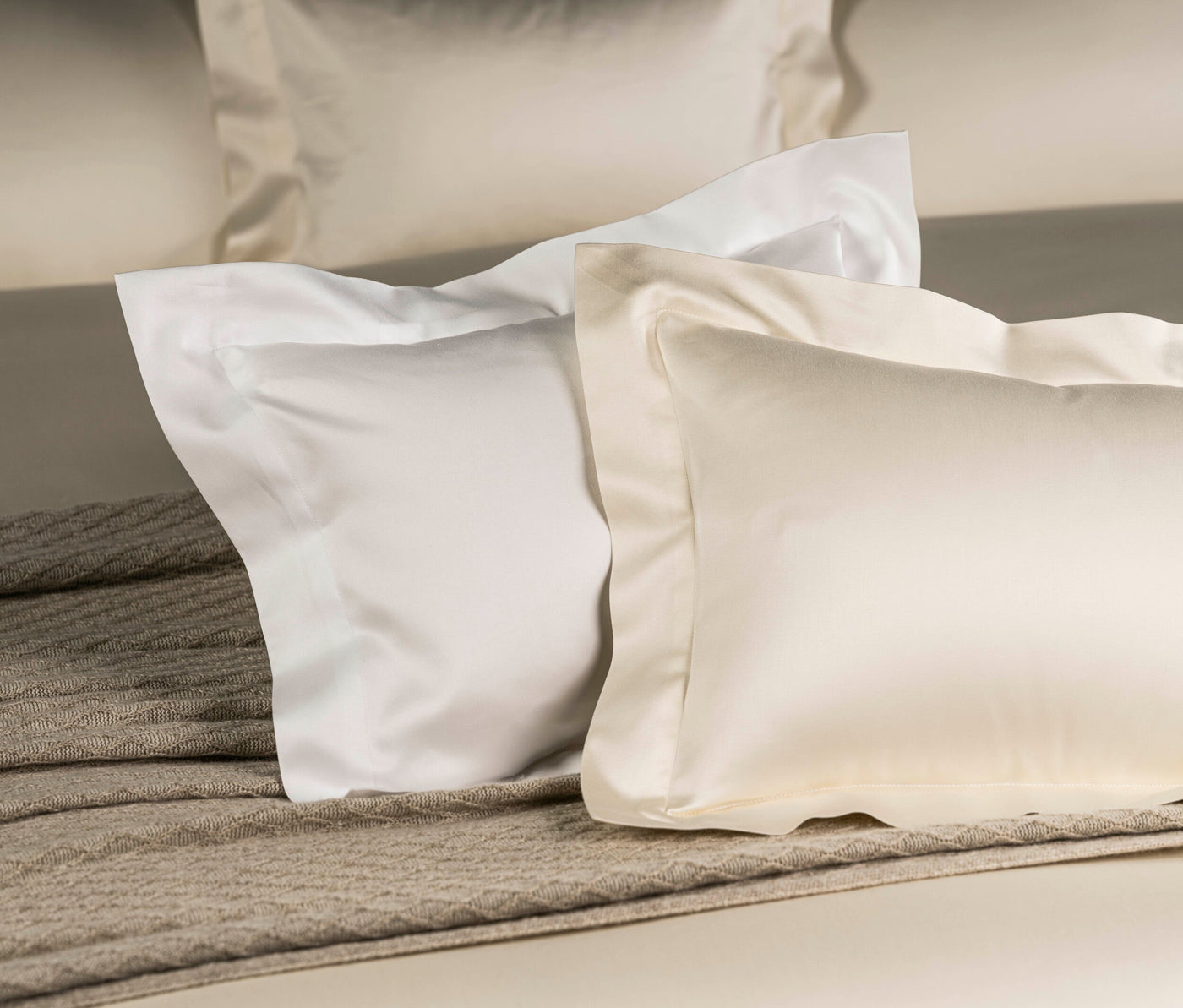 Two pillowcases, one white and one cream, are layered on a bed with a textured throw. featuring Capela Pillowcase by Celso de Lemos available at Linen Society.
