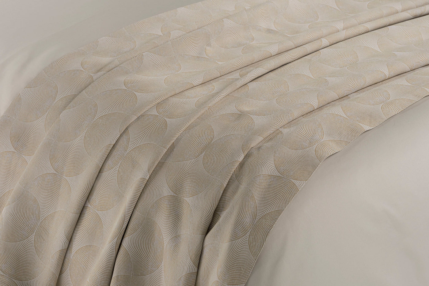 A textured, light beige bed cover with a subtle, repeated geometric fan pattern is draped across a bed. featuring Caviar Bed Cover by Celso de Lemos available at Linen Society.