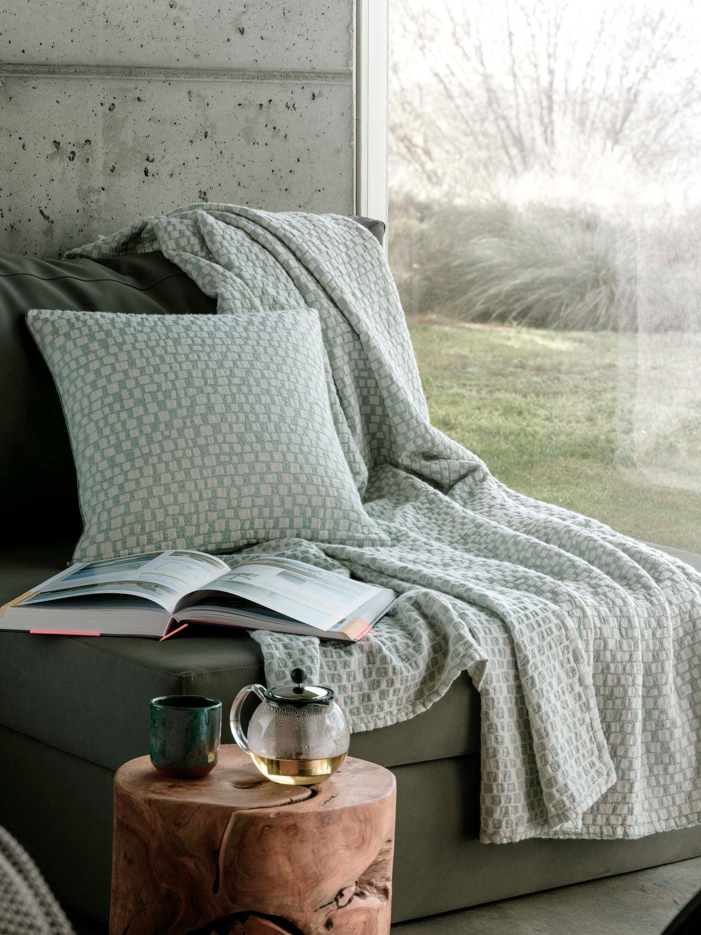 A textured teal and white woven pillow cover rests on a dark green sofa next to an open book and a tea set. featuring City Pillow Cover by Celso de Lemos available at Linen Society.