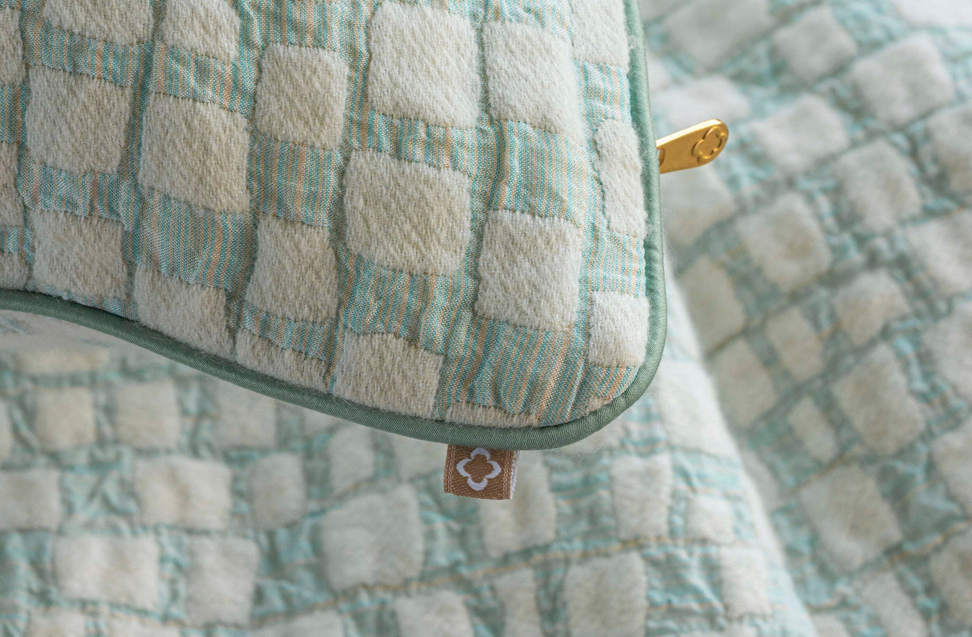 A close-up shows a textured pillow with a woven checkered pattern in cream, light green, and subtle gold threads, featuring the City Pillow Cover by Celso de Lemos available at Linen Society.