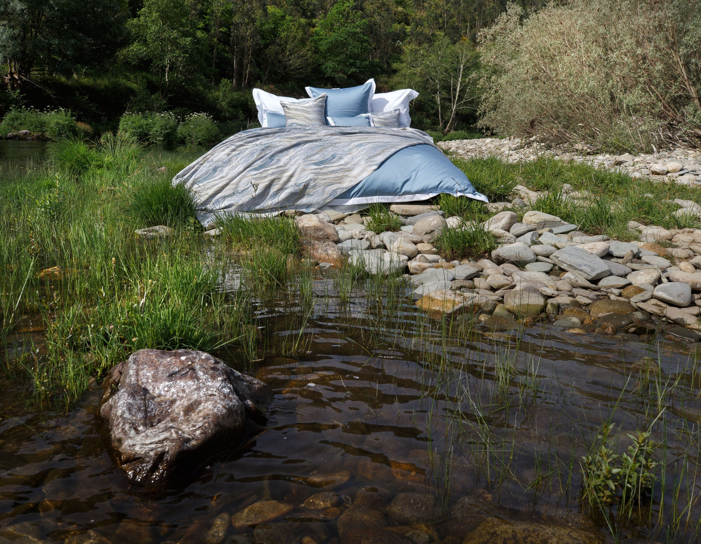 A bed with light blue and abstract patterned linens is displayed outdoors by a rocky stream. featuring Emma Pillowcase by Celso de Lemos available at Linen Society.