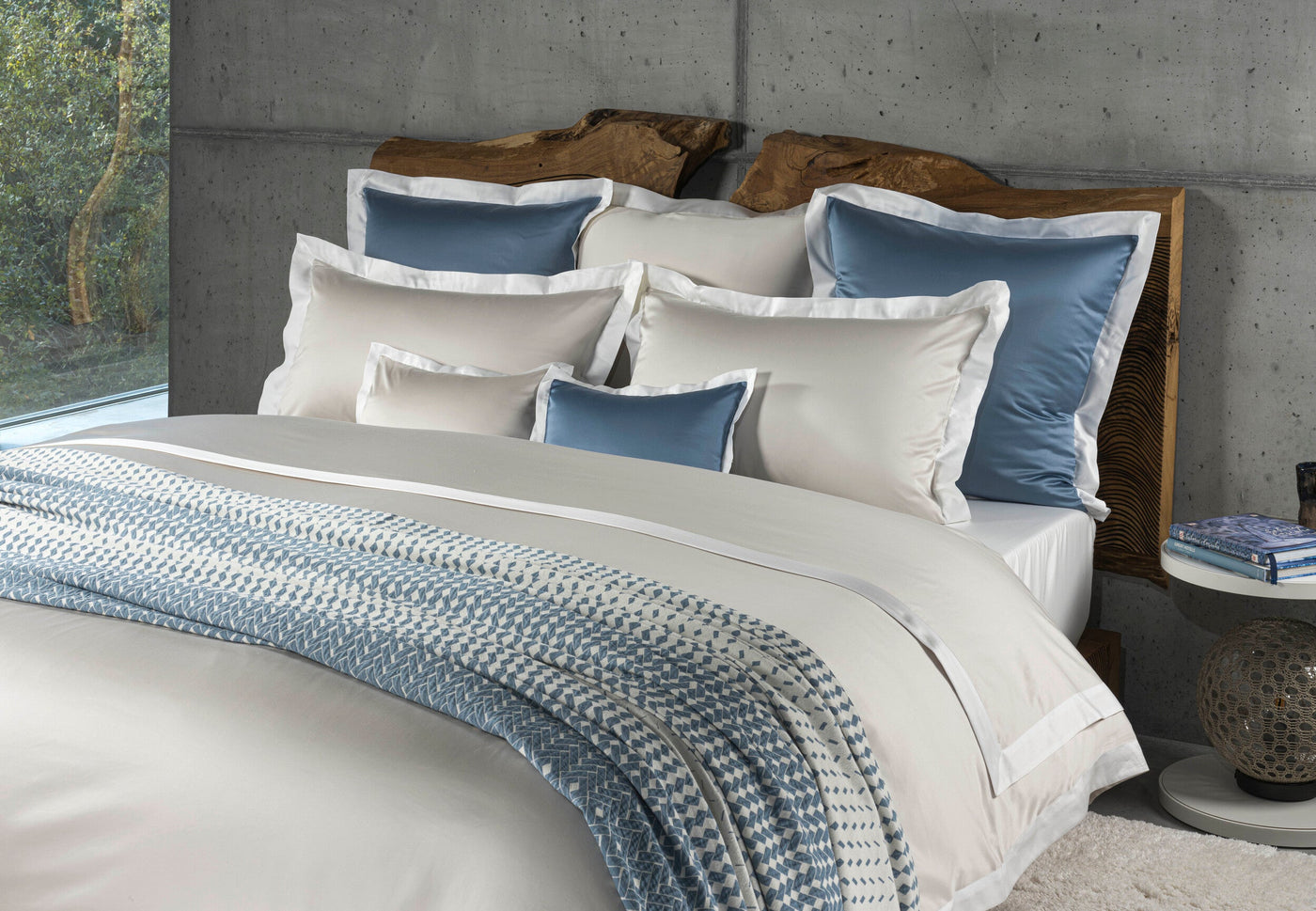 A bedroom with a bed dressed in neutral-toned linens, accented with blue pillows and a patterned throw, featuring Emma Pillowcase by Celso de Lemos available at Linen Society.