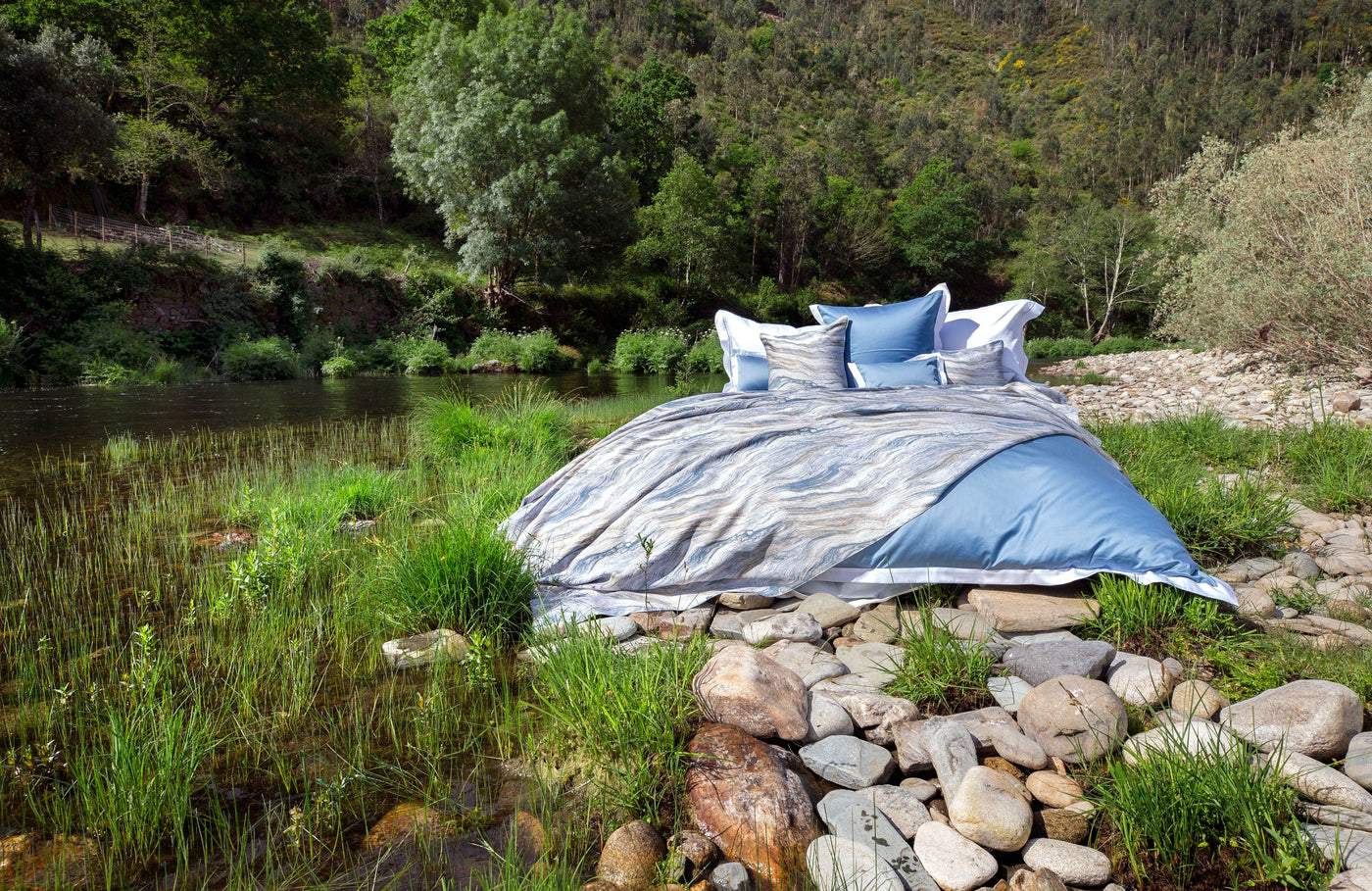 Bedding arranged on river rocks with a forest and water backdrop, featuring the Emma Pillowcase by Celso de Lemos available at Linen Society.