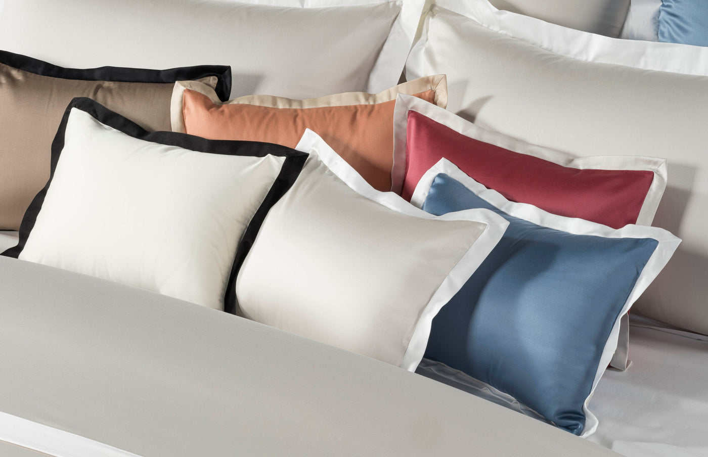 A layered display of multiple pillowcases in white, taupe, coral, red, and blue, some with contrasting black or white trim. featuring Emma Pillowcase by Celso de Lemos available at Linen Society.