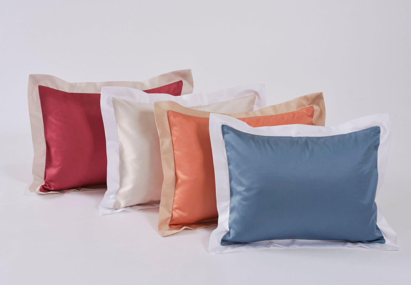 Four satin pillowcases in red, cream, coral, and blue are displayed in a staggered row, featuring Emma Pillowcase by Celso de Lemos available at Linen Society.