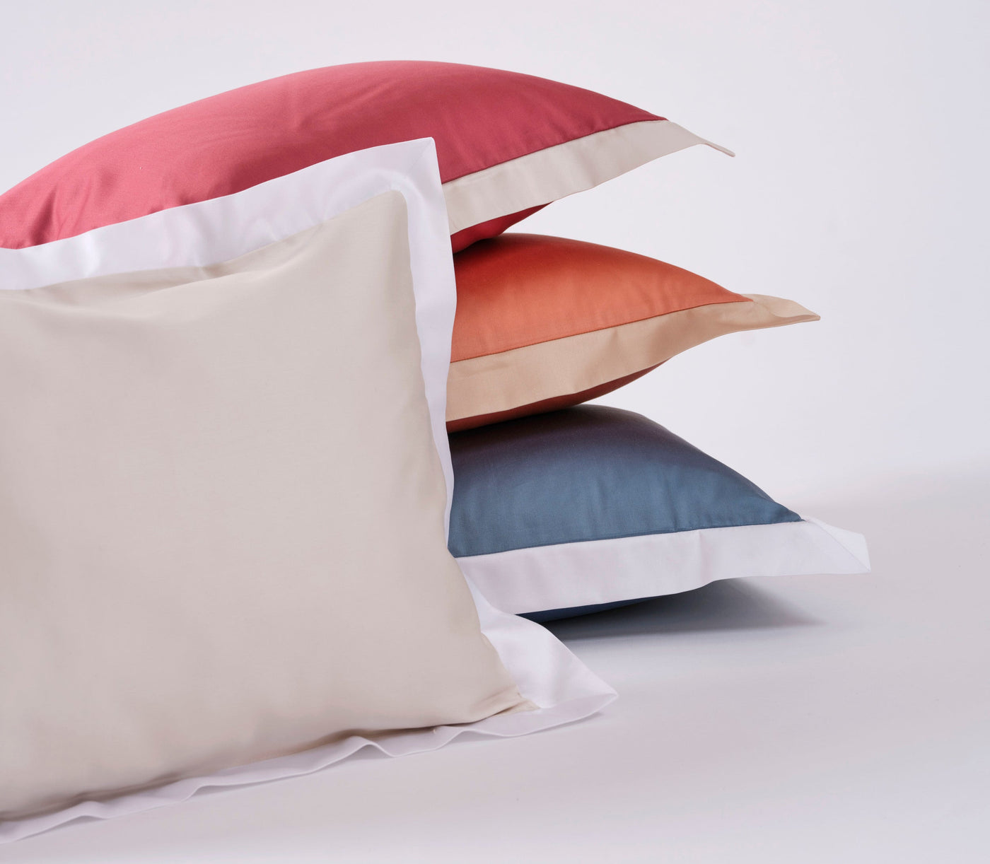 Three stacked pillowcases in beige, terracotta, and blue, each with white trim, featuring the Emma Pillowcase by Celso de Lemos available at Linen Society.