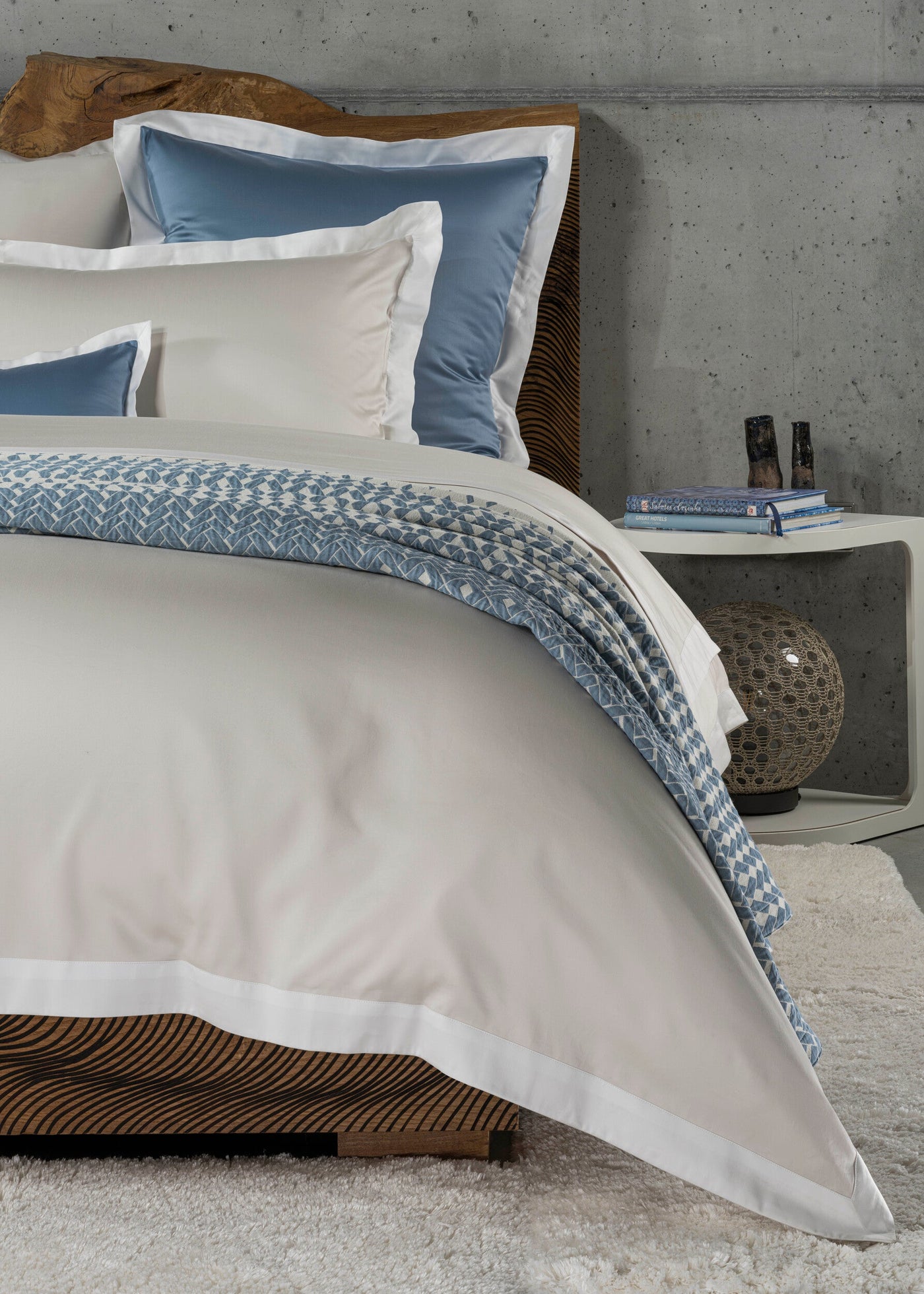 A beige duvet and pillows with blue accents are styled on a bed with a wooden headboard and a textured throw, featuring Emma Pillowcase by Celso de Lemos available at Linen Society.