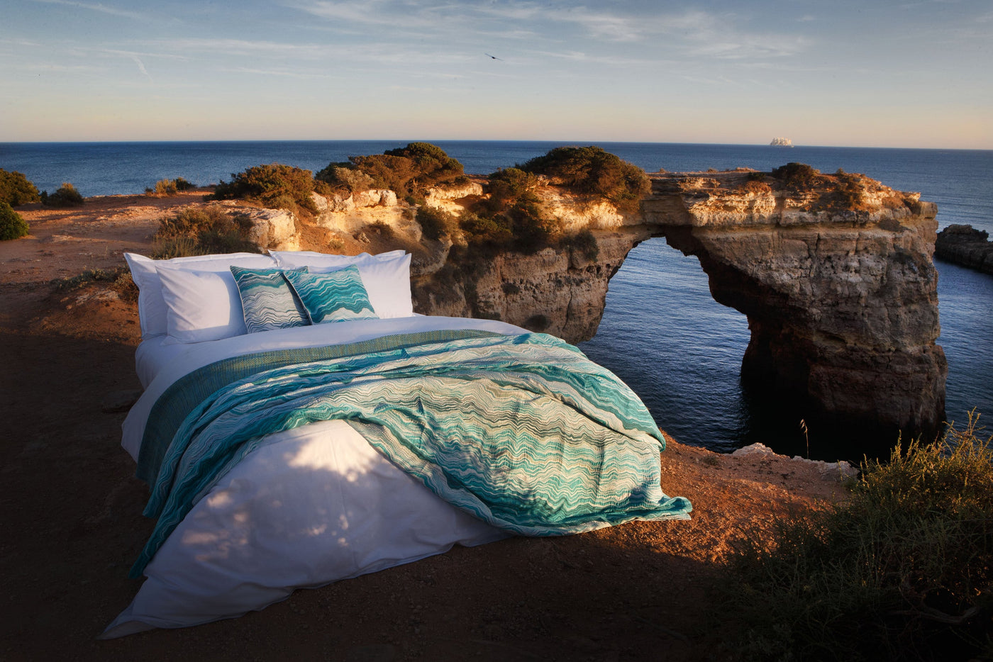 A bed dressed in white linens and a turquoise patterned throw is placed on a cliff overlooking the ocean, featuring Estrela Pillowcase by Celso de Lemos available at Linen Society.