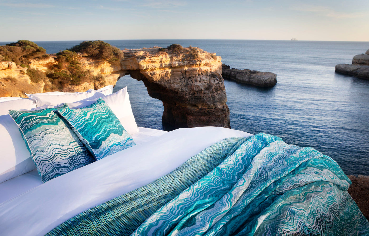 A bed with white linens and turquoise patterned pillows sits on a cliff overlooking the ocean, featuring Estrela Pillowcase by Celso de Lemos available at Linen Society.