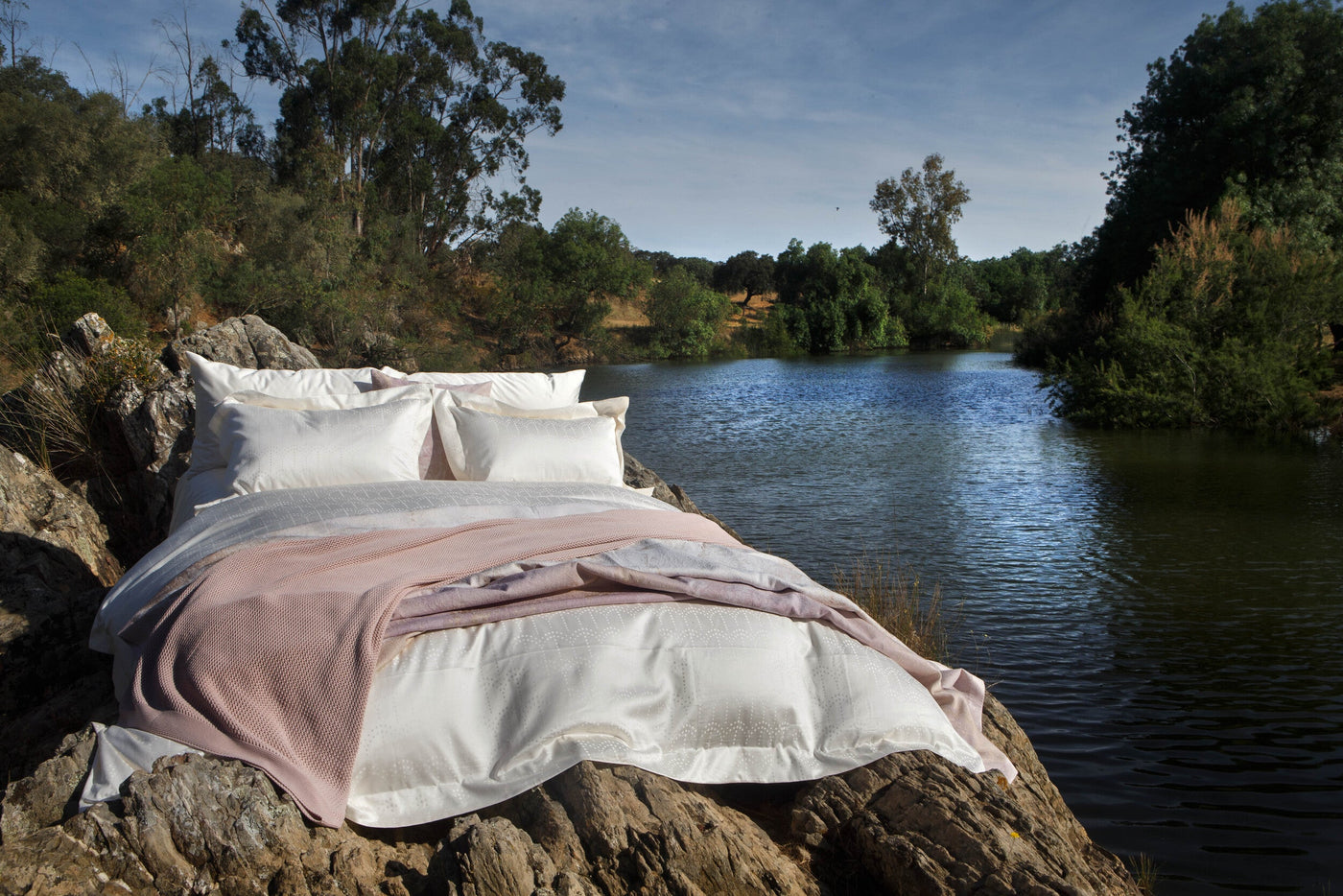 White pillowcases with subtle patterned texture are layered on a rocky outcrop next to a lake, featuring Evora Pillowcase by Celso de Lemos available at Linen Society.