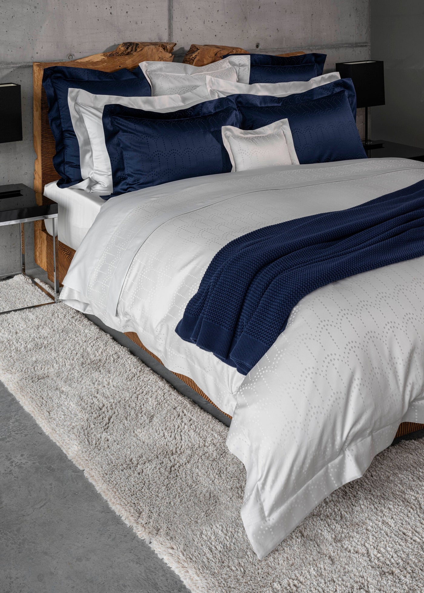 A bed with white bedding and navy blue accent pillows and throw. featuring Evora Pillowcase by Celso de Lemos available at Linen Society.