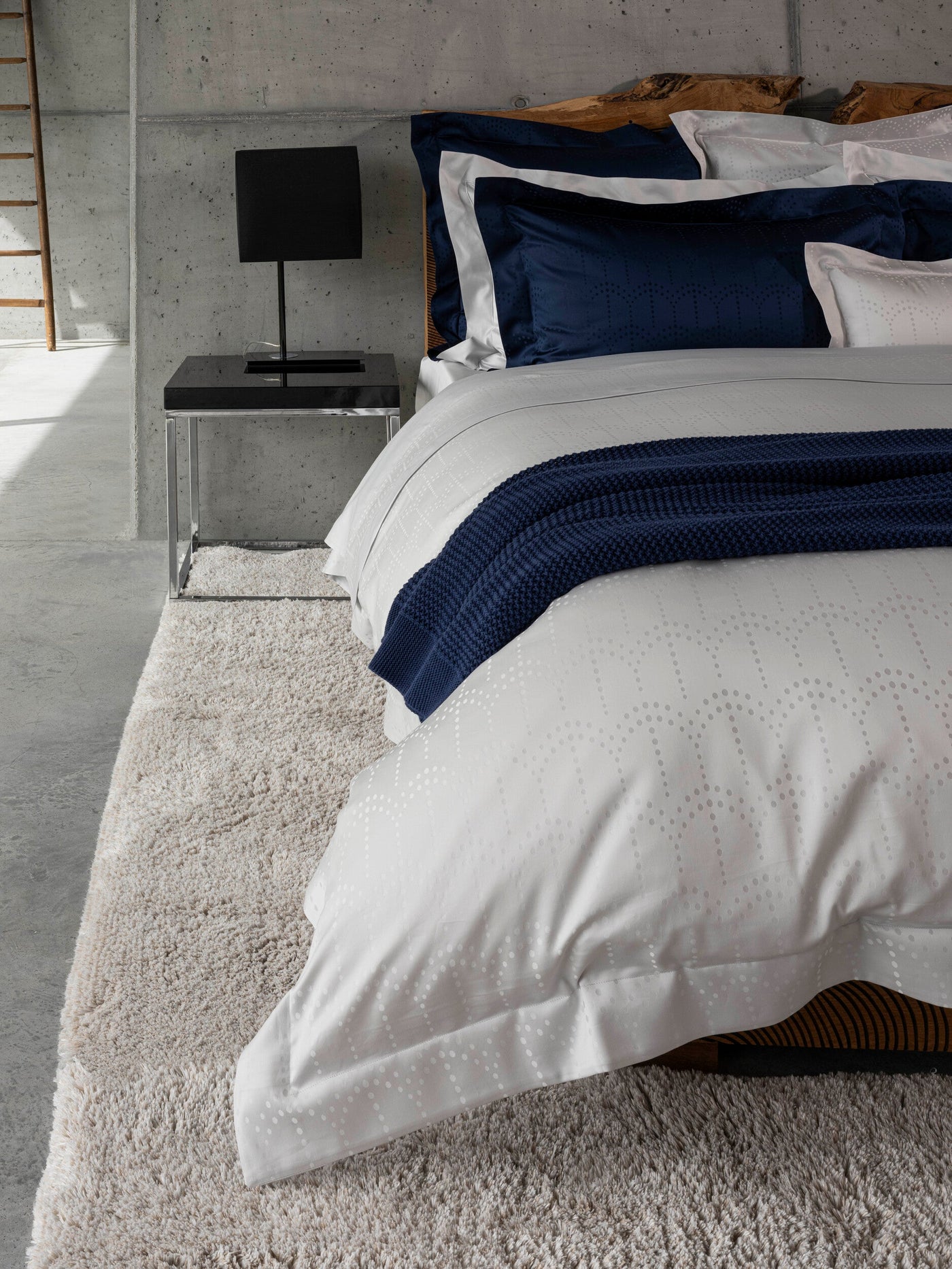 A white Pillowcase with a subtle dotted pattern is layered on a bed with navy blue accents and a textured rug, featuring Evora Pillowcase by Celso de Lemos available at Linen Society.