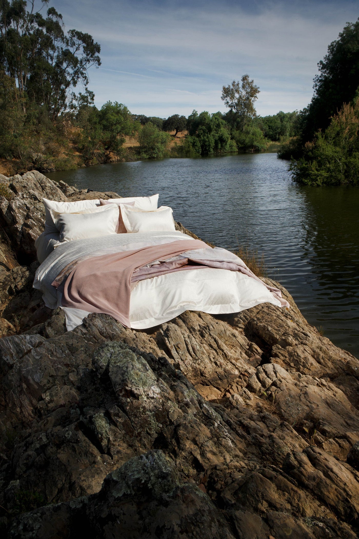 White bedding and a blush throw are arranged on rocky terrain by a calm body of water, featuring Evora Pillowcase by Celso de Lemos available at Linen Society.