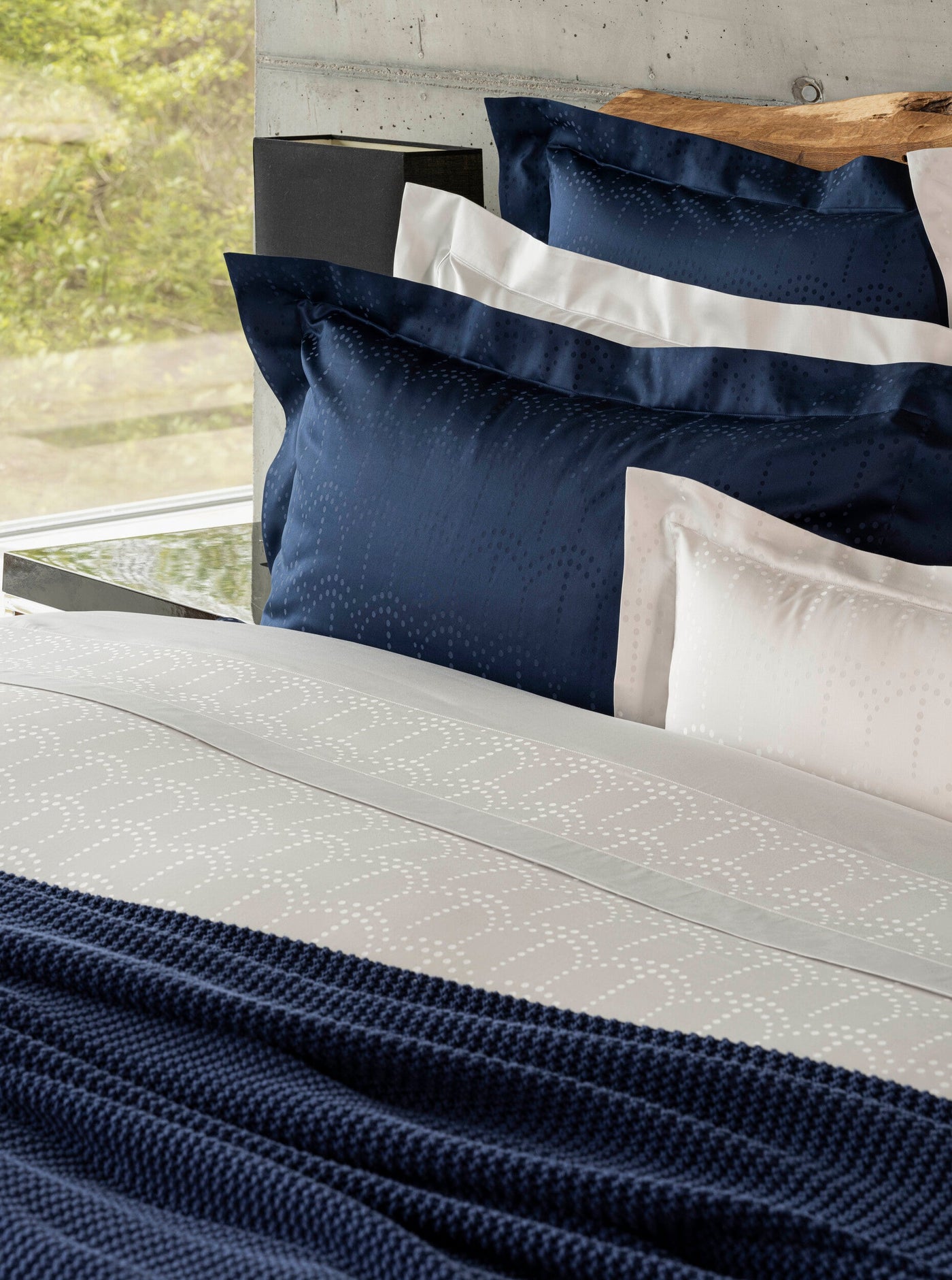 A bed with navy blue and white patterned bedding, a navy knitted throw, and a wooden headboard, featuring Evora Pillowcase by Celso de Lemos available at Linen Society.