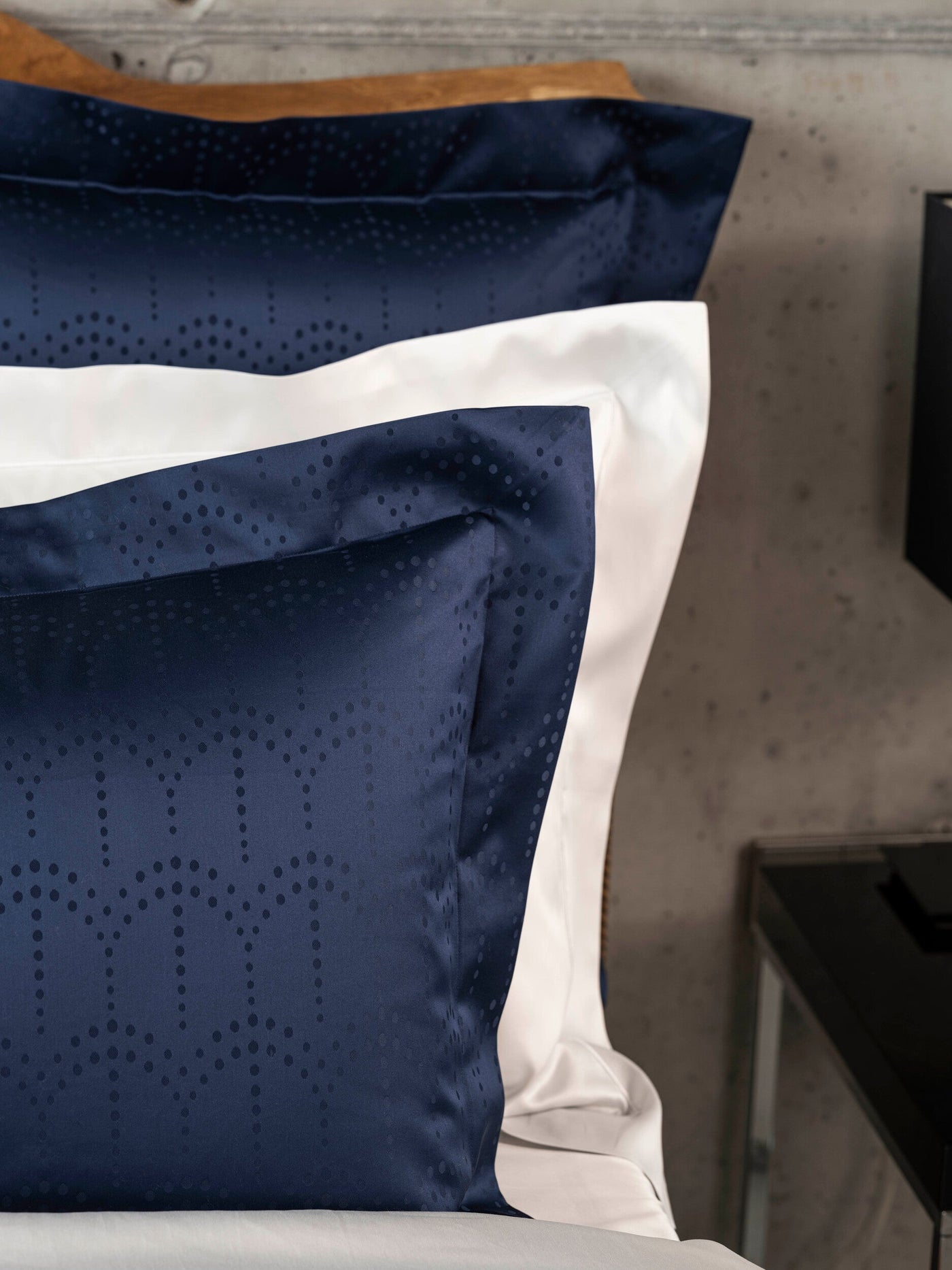 Close-up of dark navy pillowcases with a subtle dot pattern layered with white linens, featuring Evora Pillowcase by Celso de Lemos available at Linen Society.