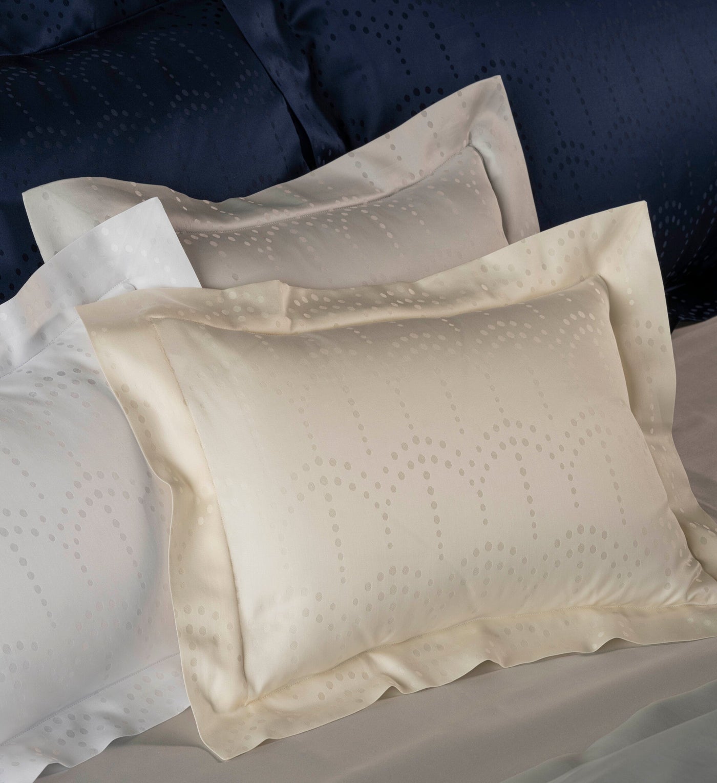 Cream, white, and navy pillowcases with a subtle dot pattern are arranged on a bed, featuring Evora Pillowcase by Celso de Lemos available at Linen Society.