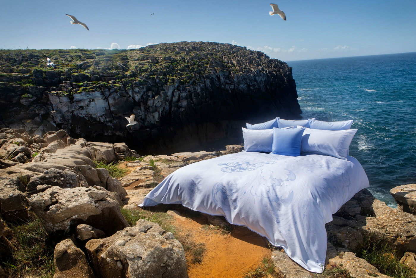 A bed with light blue bedding and intricate floral patterns is set up on a rocky coastline with the ocean in the background. featuring Exquise Pillowcase by Celso de Lemos available at Linen Society.