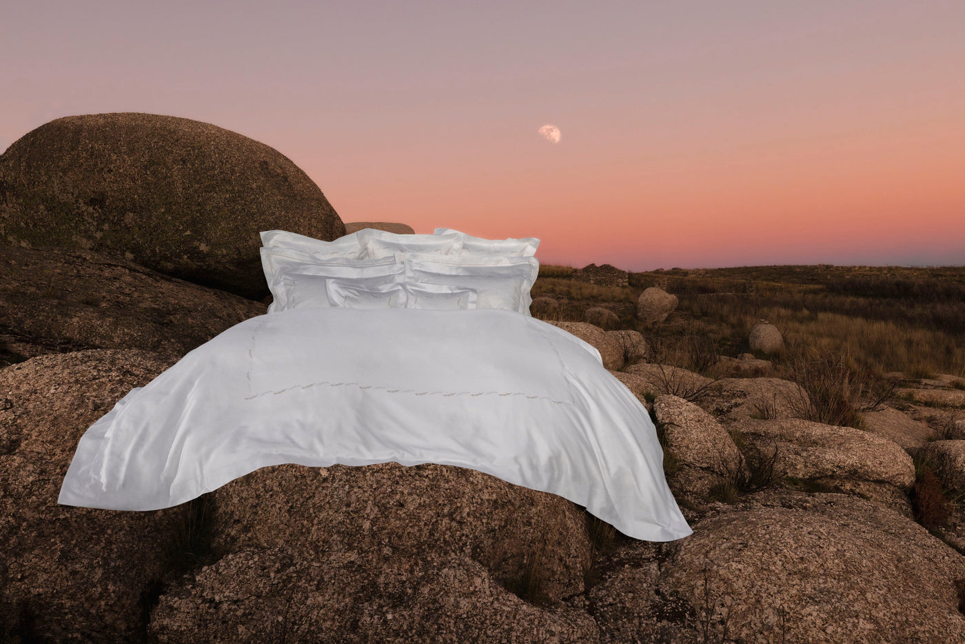 White linen bedding is artfully draped on large rocks with a soft, gradient sunset sky in the background, featuring Fern Pillowcase by Celso de Lemos available at Linen Society.
