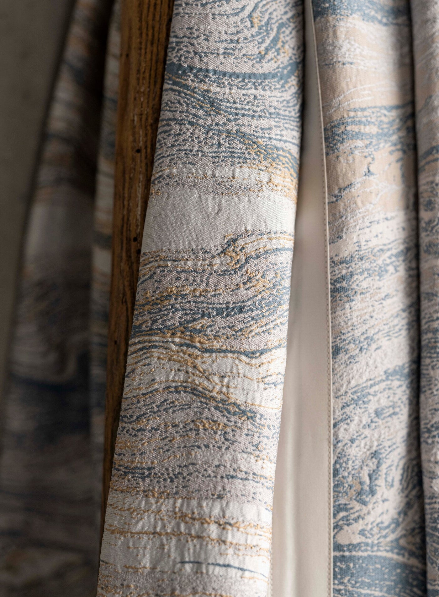 Close-up of a textured, woven fabric with a marbled pattern in shades of blue, cream, and gold, draped over a wooden surface. featuring Filigrane Bed Cover by Celso de Lemos available at Linen Society.