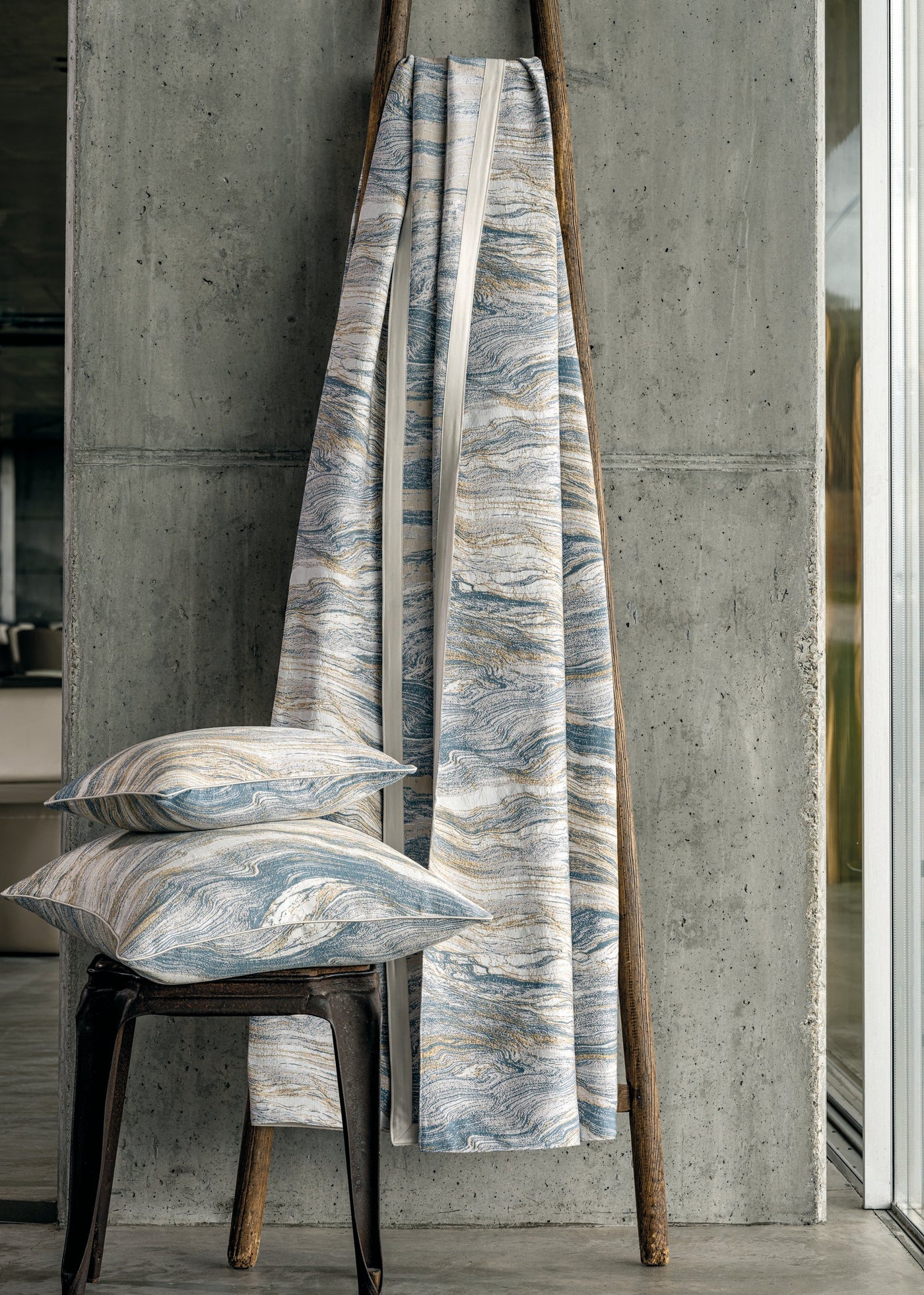 Two patterned pillows are stacked on a dark metal stool next to a draped textile on a wooden ladder, featuring the Filigrane Pillow Cover by Celso de Lemos available at Linen Society.