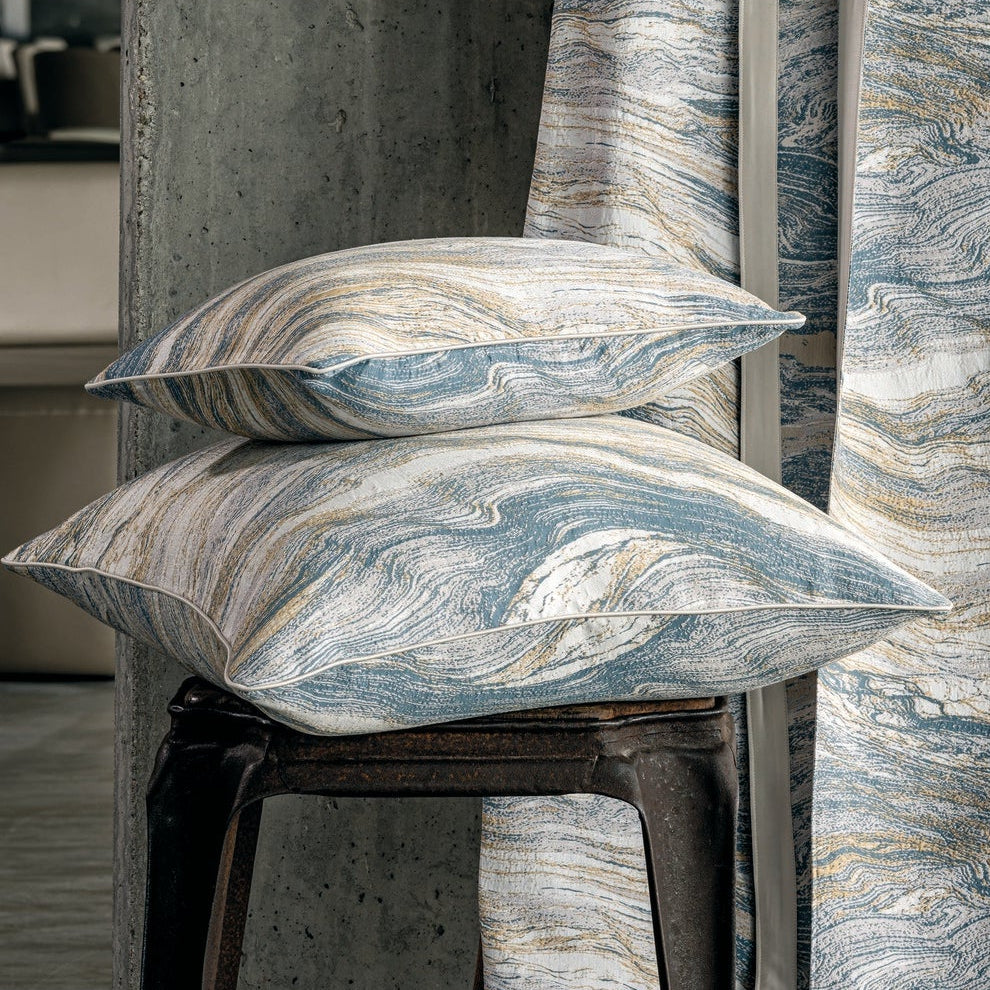 Two patterned pillows are stacked on a dark metal stool next to a draped textile on a wooden ladder, featuring the Filigrane Pillow Cover by Celso de Lemos available at Linen Society.