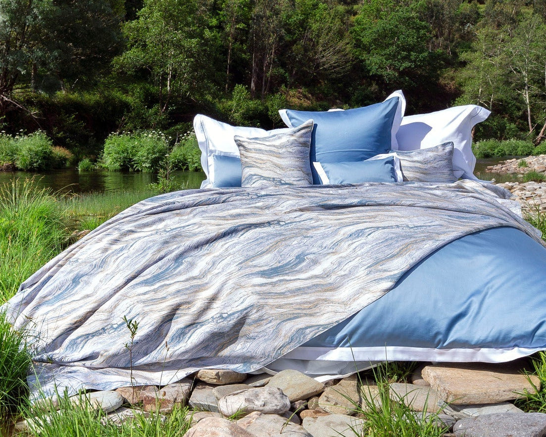 A bed adorned with a marble-patterned duvet and blue accents sits outdoors beside a river. featuring Filigrane Pillow Cover by Celso de Lemos available at Linen Society.