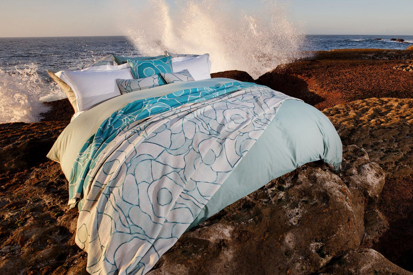 A bed with white and aqua patterned bedding is set against a rocky coastline with waves crashing. featuring Funchal Pillow Cover by Celso de Lemos available at Linen Society.