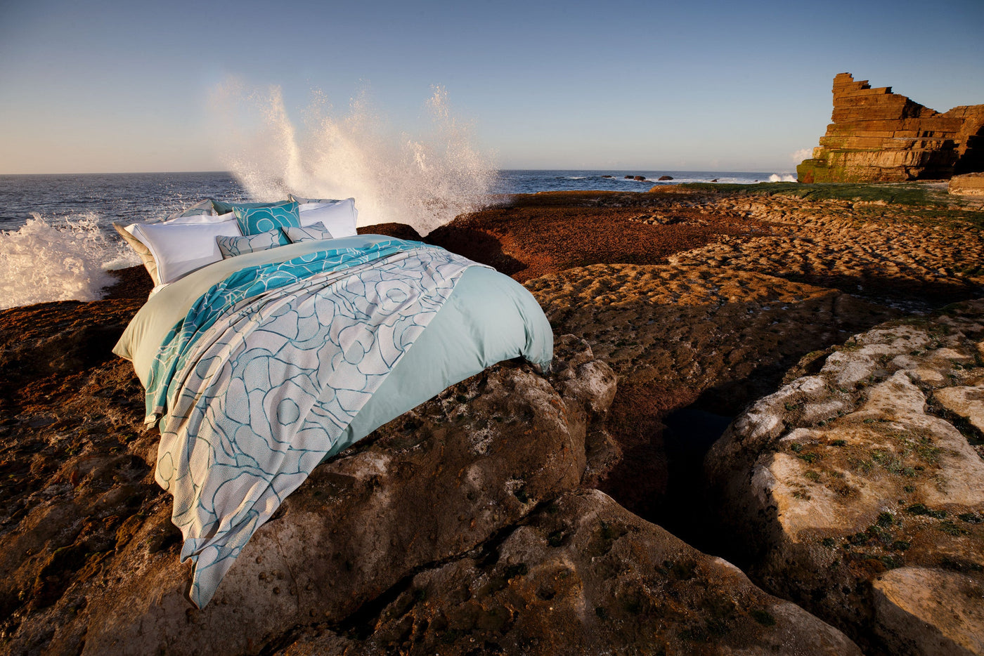 A bed with white and aqua patterned bedding is set against a rocky coastline with waves crashing. featuring Funchal Pillow Cover by Celso de Lemos available at Linen Society.