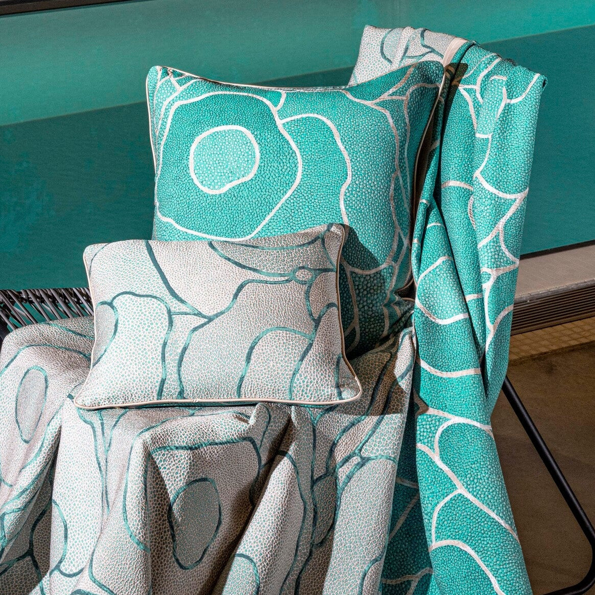 A chair draped with teal and grey patterned cushions and a throw blanket by a pool featuring Funchal Pillow Cover by Celso de Lemos available at Linen Society.