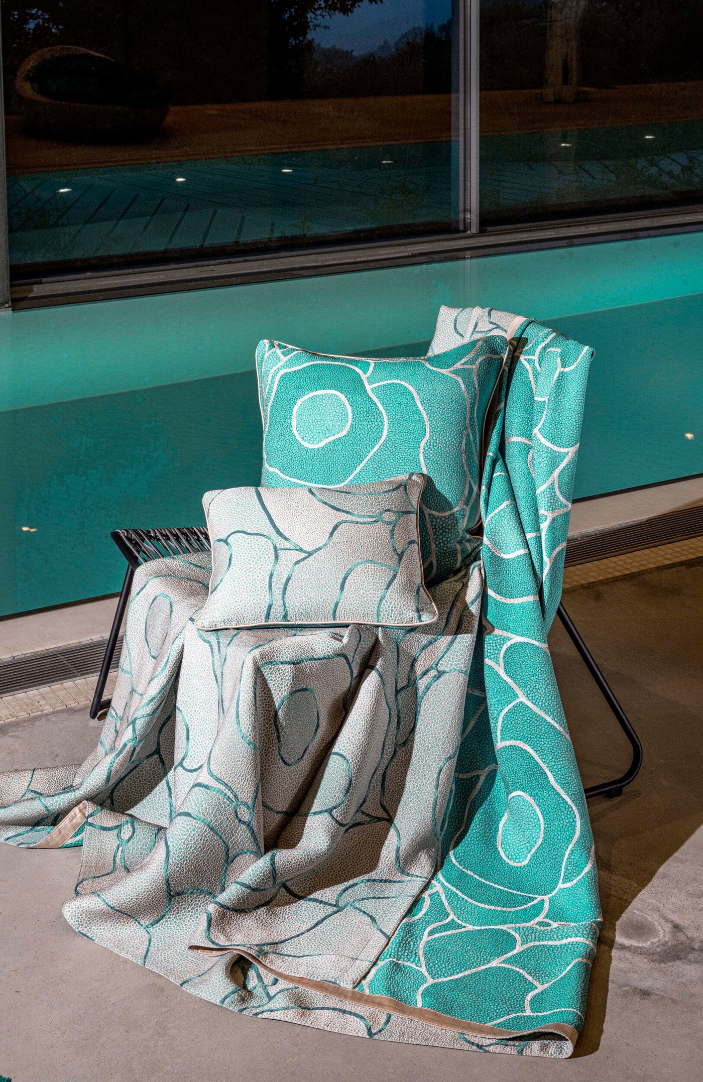 A chair draped with teal and grey patterned cushions and a throw blanket by a pool featuring Funchal Pillow Cover by Celso de Lemos available at Linen Society.