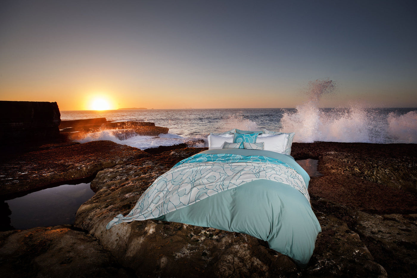 A bed with a teal and white patterned duvet is set on rocky terrain by the ocean at sunset, featuring the Funchal Pillow Cover by Celso de Lemos available at Linen Society.