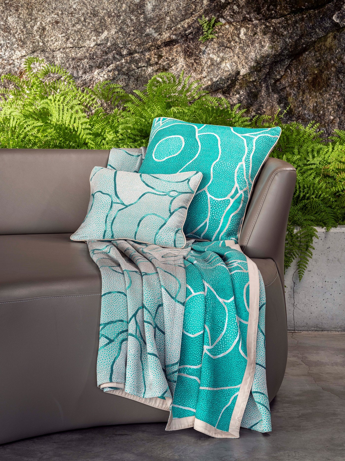 Two aqua and white patterned pillow covers and a matching throw blanket are displayed on a dark grey sofa, with ferns and a textured rock wall in the background, featuring Funchal Pillow Cover by Celso de Lemos available at Linen Society.
