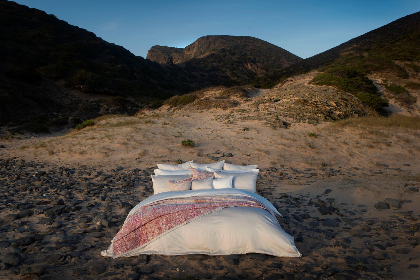 A neatly made bed with white linens and a textured pink and white throw blanket is set against a backdrop of sand dunes and mountains. featuring Gemini Pillowcase by Celso de Lemos available at Linen Society.