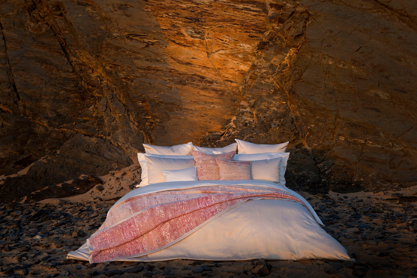 A bed with crisp white linens and textured blush accent pillows and a throw is set against a rocky, sunlit cliff face. featuring Gemini Pillowcase by Celso de Lemos available at Linen Society.