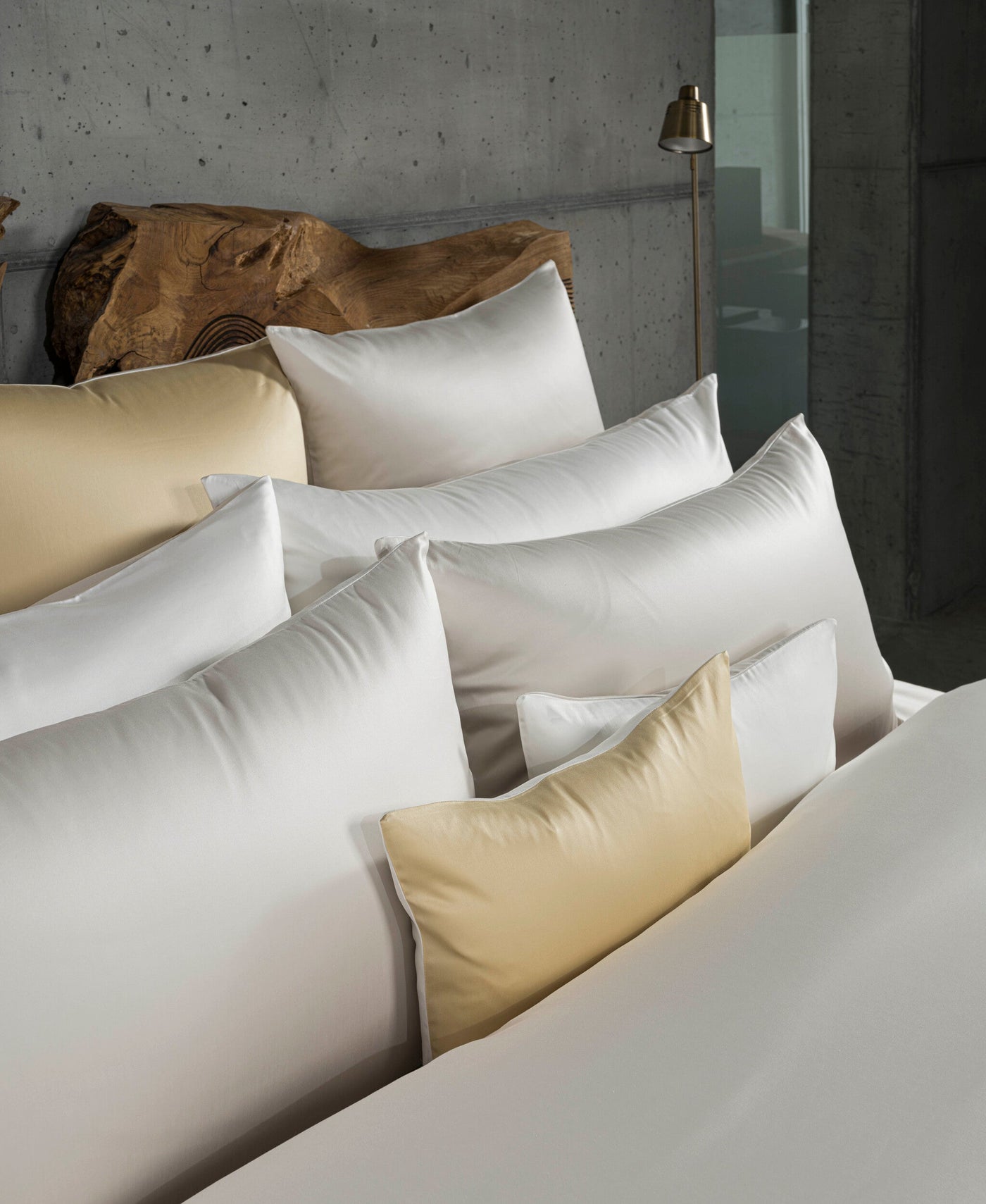 A pile of white and gold pillowcases are arranged in front of a textured wooden headboard against a concrete wall. featuring Gemini Pillowcase by Celso de Lemos available at Linen Society.