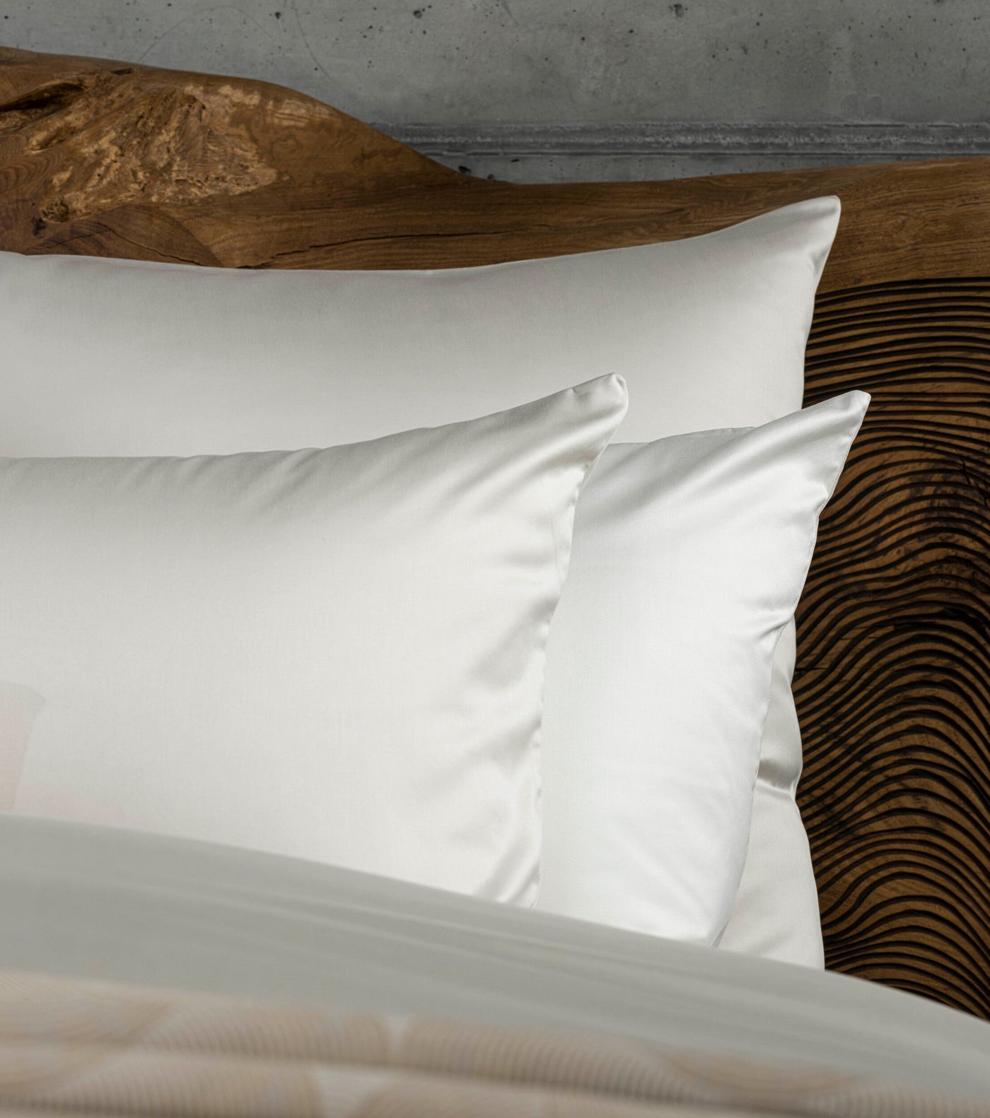 Two white silk pillowcases are neatly arranged on a bed with a textured wooden headboard, featuring Gemini Pillowcase by Celso de Lemos available at Linen Society.