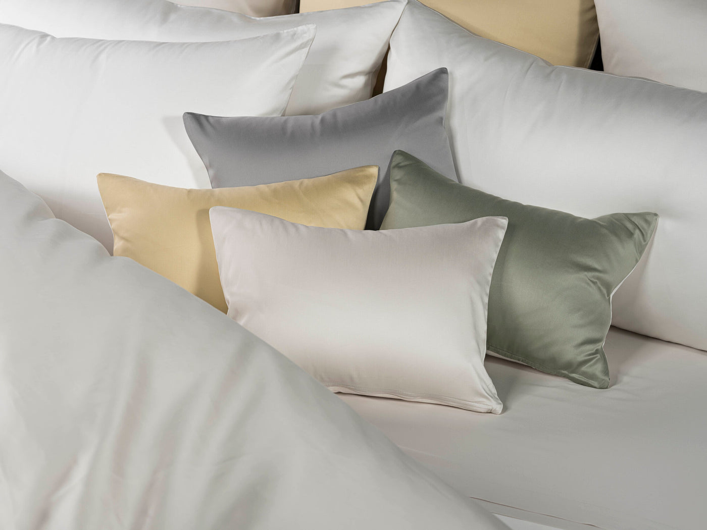 A collection of luxuriously soft pillowcases in shades of white, pale yellow, grey, and sage green, featuring Gemini Pillowcase by Celso de Lemos available at Linen Society.