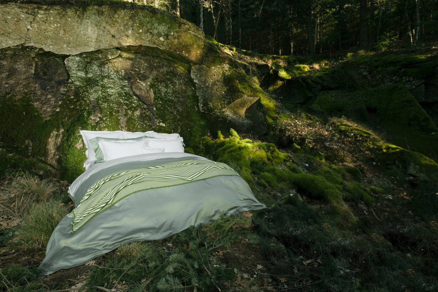 A bed with white and sage green bedding is set against mossy rocks and forest floor, featuring Ivy Pillowcase by Celso de Lemos available at Linen Society.