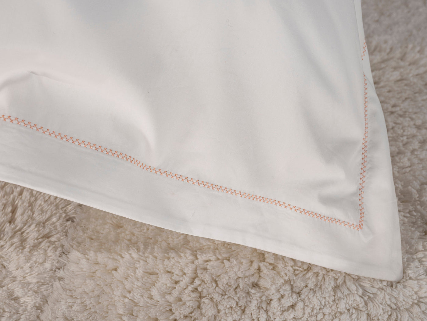 A white pillowcase with coral orange stitching on a textured beige rug, featuring Ivy Pillowcase by Celso de Lemos available at Linen Society.