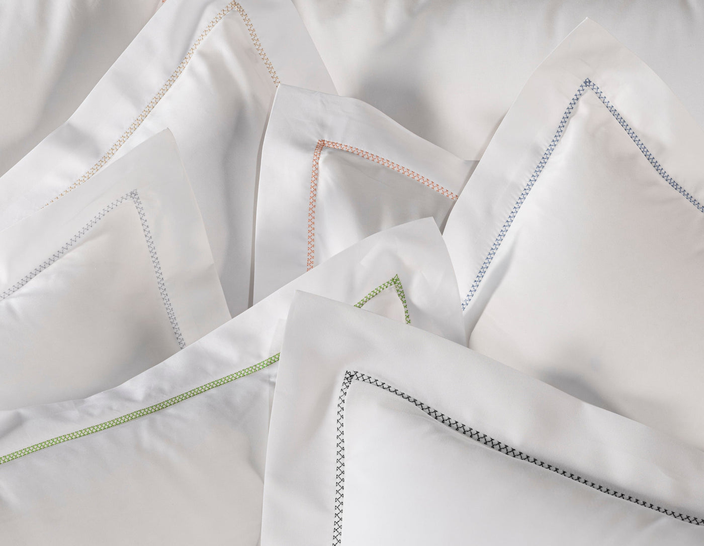 A pile of white pillowcases with a textured embroidered border in various colors like gold, coral, blue, green, and black, featuring the Ivy Pillowcase by Celso de Lemos available at Linen Society.