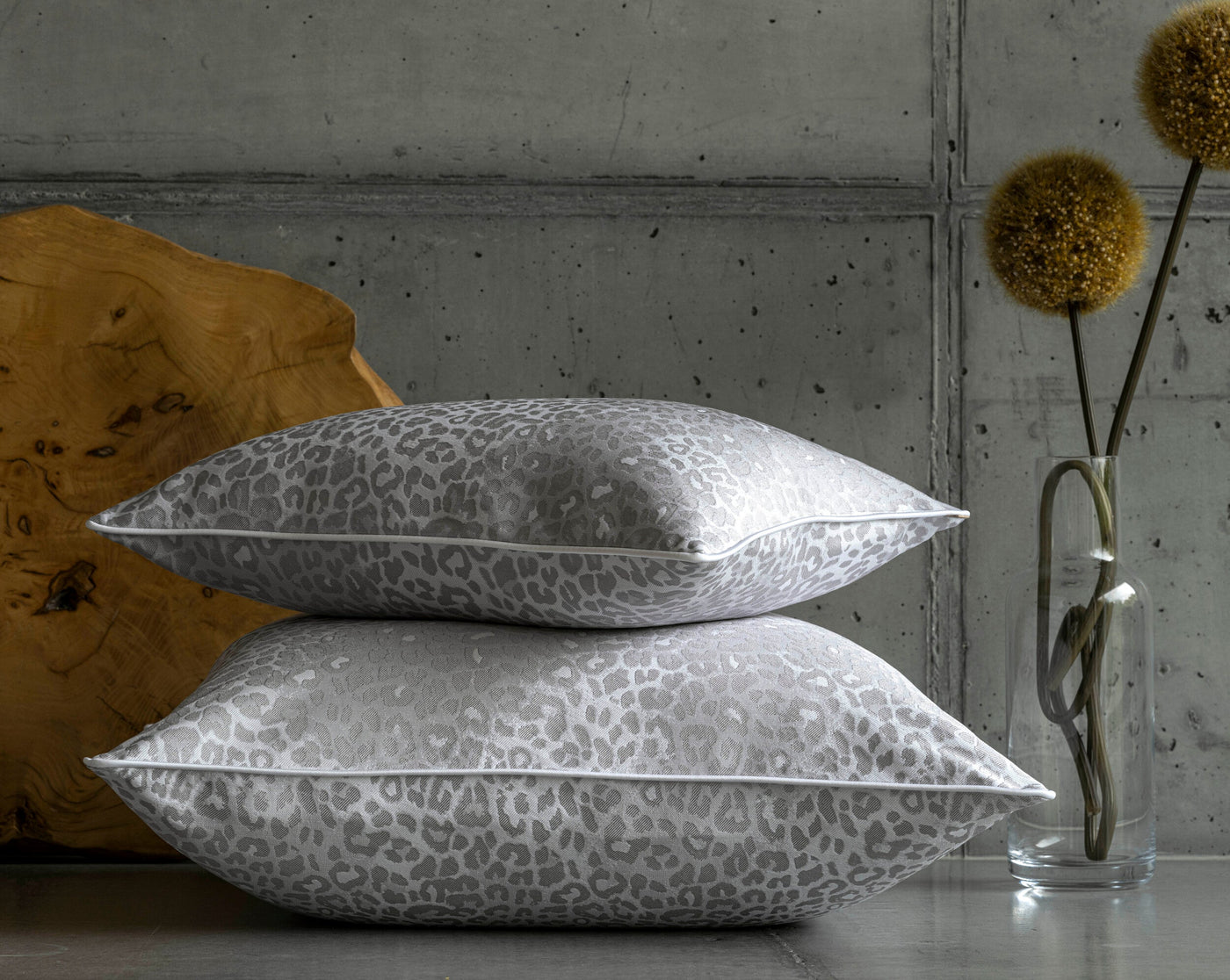 Two grey leopard print pillow covers are stacked on a surface, with a wooden accent and decorative branches in a vase in the background, featuring Jag Pillow Cover by Celso de Lemos available at Linen Society.
