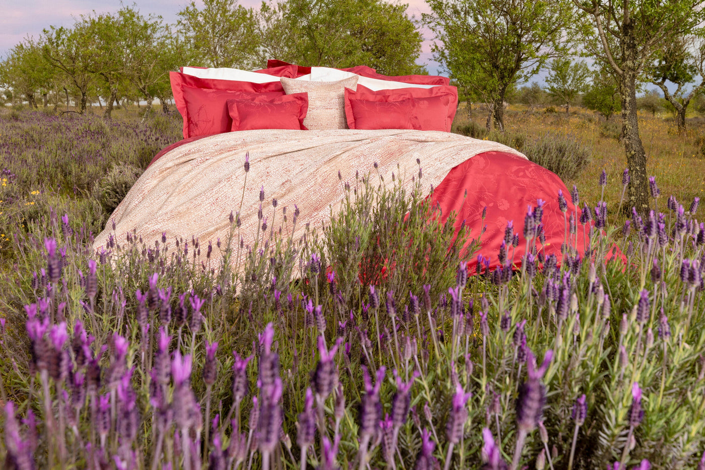 A bed with red pillows and a patterned cover is set up in a field of lavender, featuring Jardim Pillowcase by Celso de Lemos available at Linen Society.