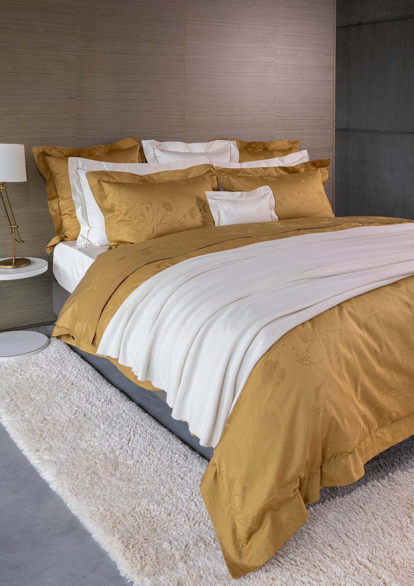 A bed dressed in golden floral patterned duvet and pillowcases, with a white knitted throw and a shaggy rug, featuring Jardim Pillowcase by Celso de Lemos available at Linen Society.