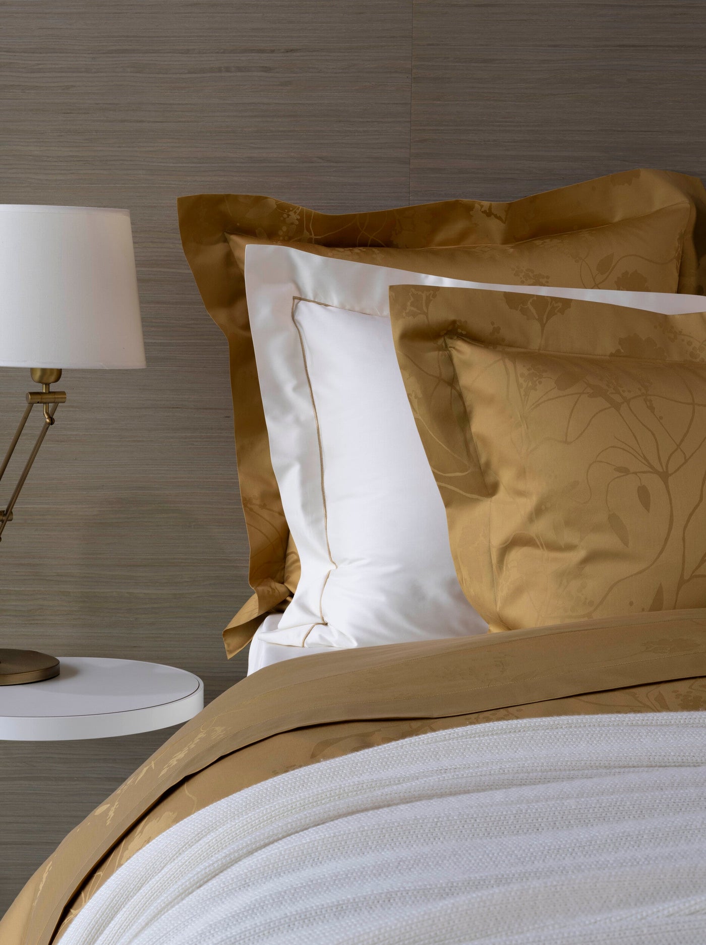 A close-up of a bed with gold embroidered pillowcases and white linens, featuring Jardim Pillowcase by Celso de Lemos available at Linen Society.