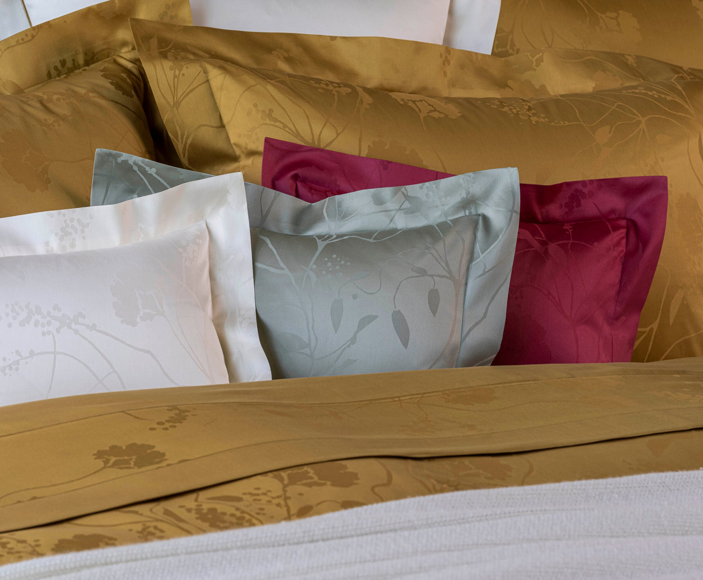 A pile of pillows in gold, white, sage green, and berry hues, each with a subtle floral pattern, featuring Jardim Pillowcase by Celso de Lemos available at Linen Society.