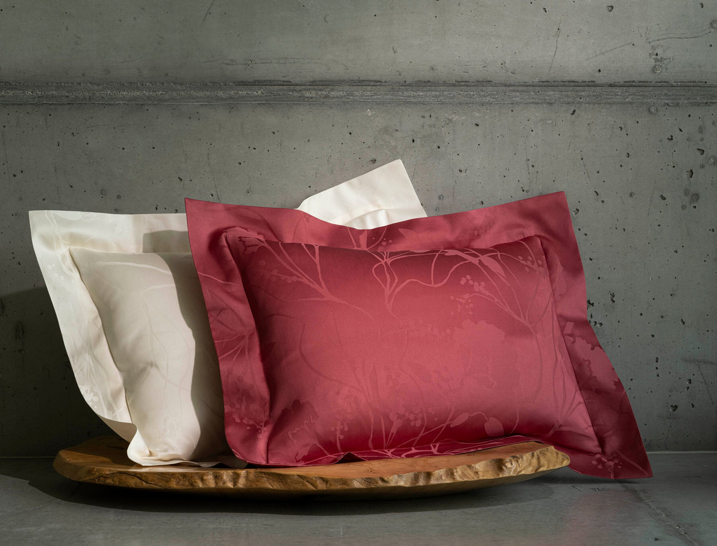 Two pillowcases, one red with a botanical pattern and one cream with a subtle design, rest on a wooden dish against a concrete backdrop. featuring Jardim Pillowcase by Celso de Lemos available at Linen Society.