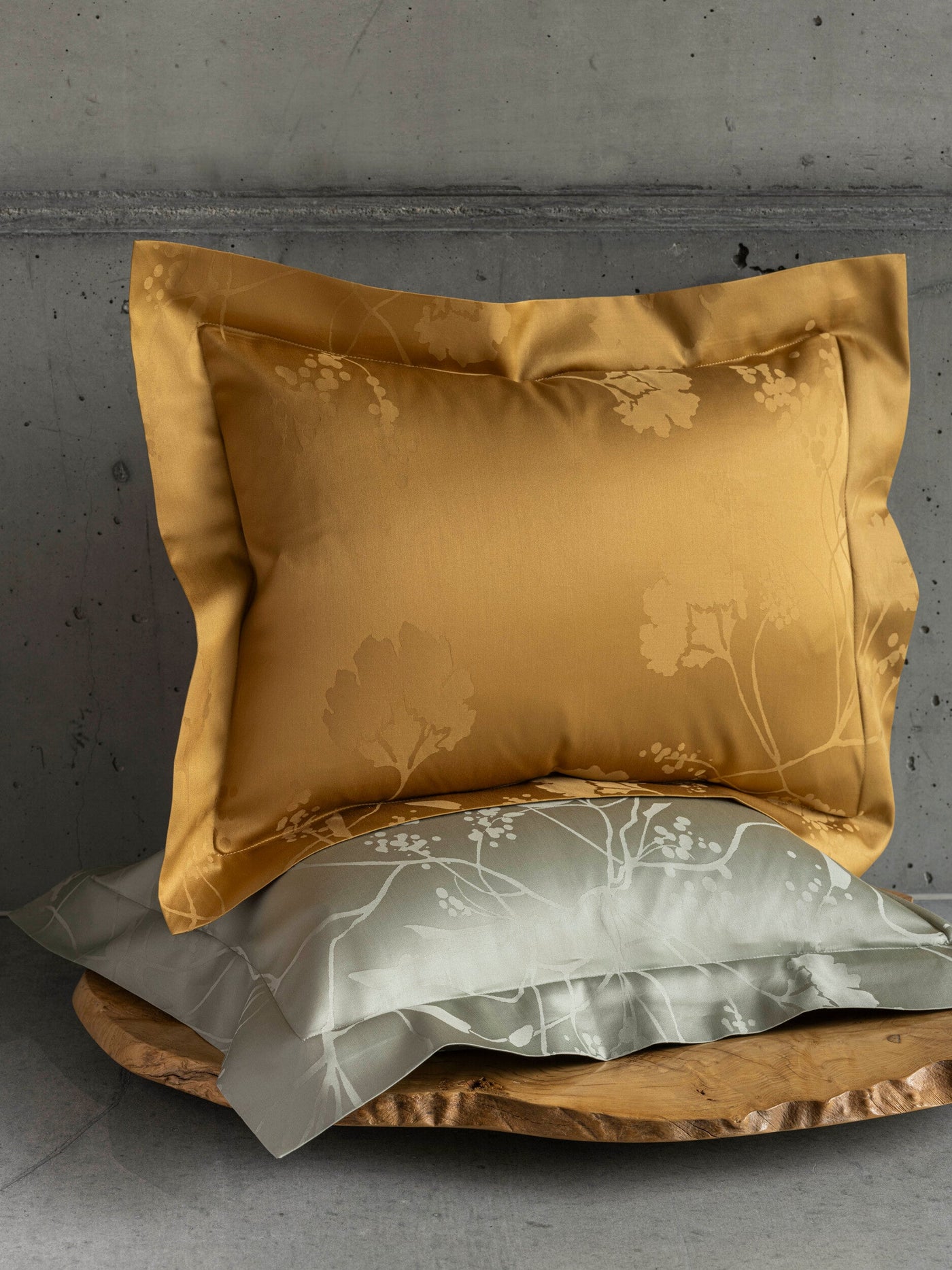 A golden yellow pillowcase with a tonal floral pattern rests atop a sage green pillowcase with a white botanical print, displayed on a rustic wooden slab against a concrete backdrop. featuring Jardim Pillowcase by Celso de Lemos available at Linen Society.