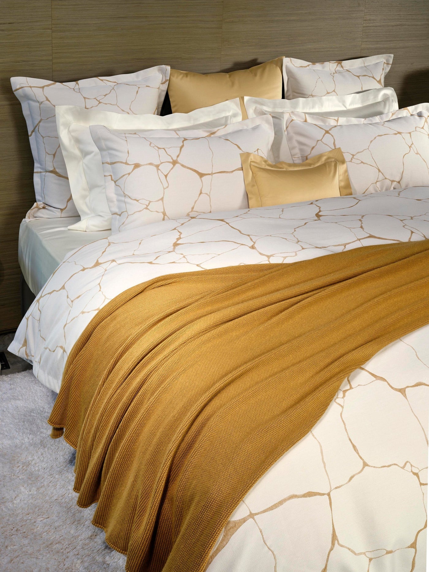 A bed with white bedding featuring a gold abstract pattern and a mustard yellow knit throw draped across it, featuring Jessie Pillowcase by Celso de Lemos available at Linen Society.
