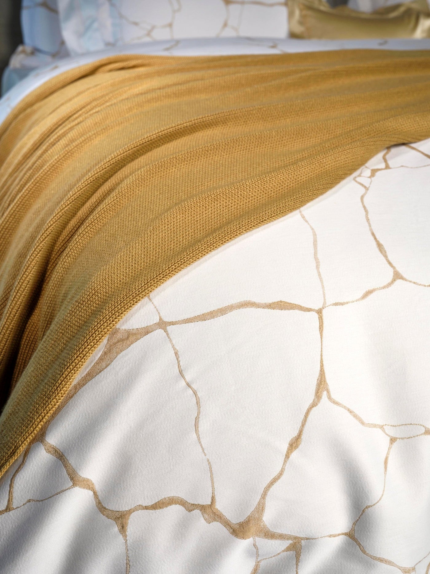 A gold knit throw covers a white pillowcase with a subtle gold abstract pattern. featuring Jessie Pillowcase by Celso de Lemos available at Linen Society.